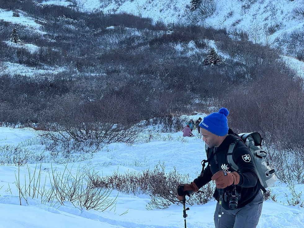 Man hiking in snowy landscape, wearing blue beanie and backpack, using trekking poles. Snowy hills and bushes in background, cold atmosphere.