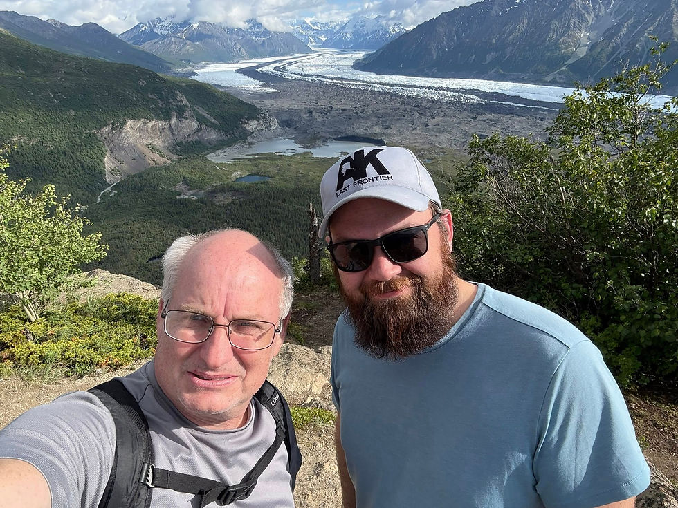 Two men smile in front of a scenic mountain view with a glacier under a blue sky. One wears a gray shirt and backpack, the other a hat and sunglasses.