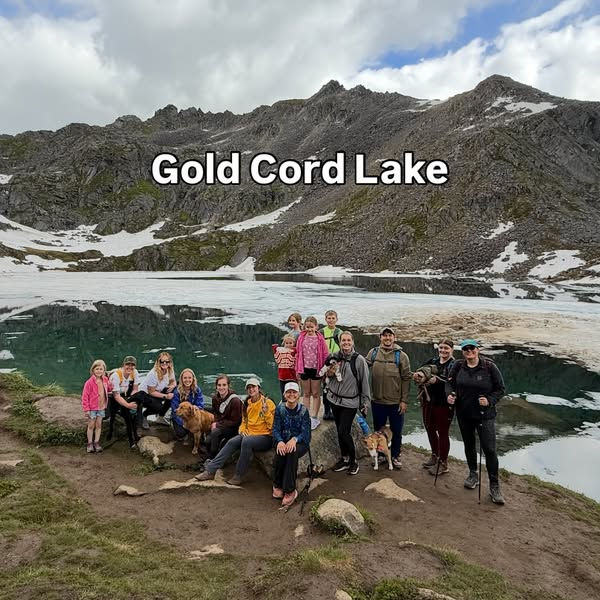 Group of people and dogs posing by Gold Cord Lake, surrounded by snowy mountains under a cloudy sky. Text reads "Gold Cord Lake."