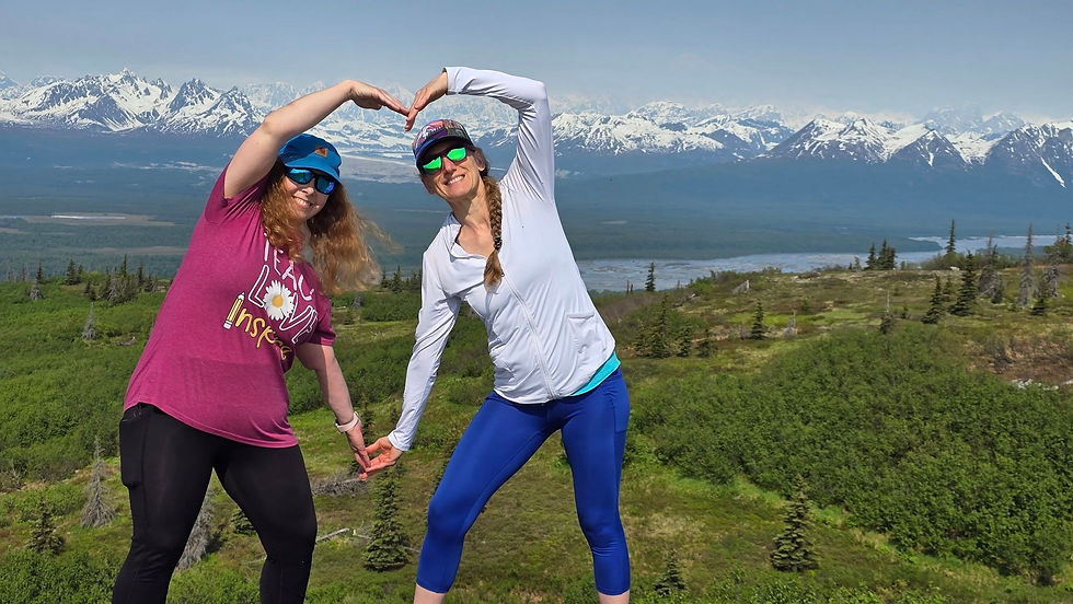 Two women form a heart shape with their arms, smiling in front of snowy mountains and green hills. One wears a red shirt; the other, a white jacket.