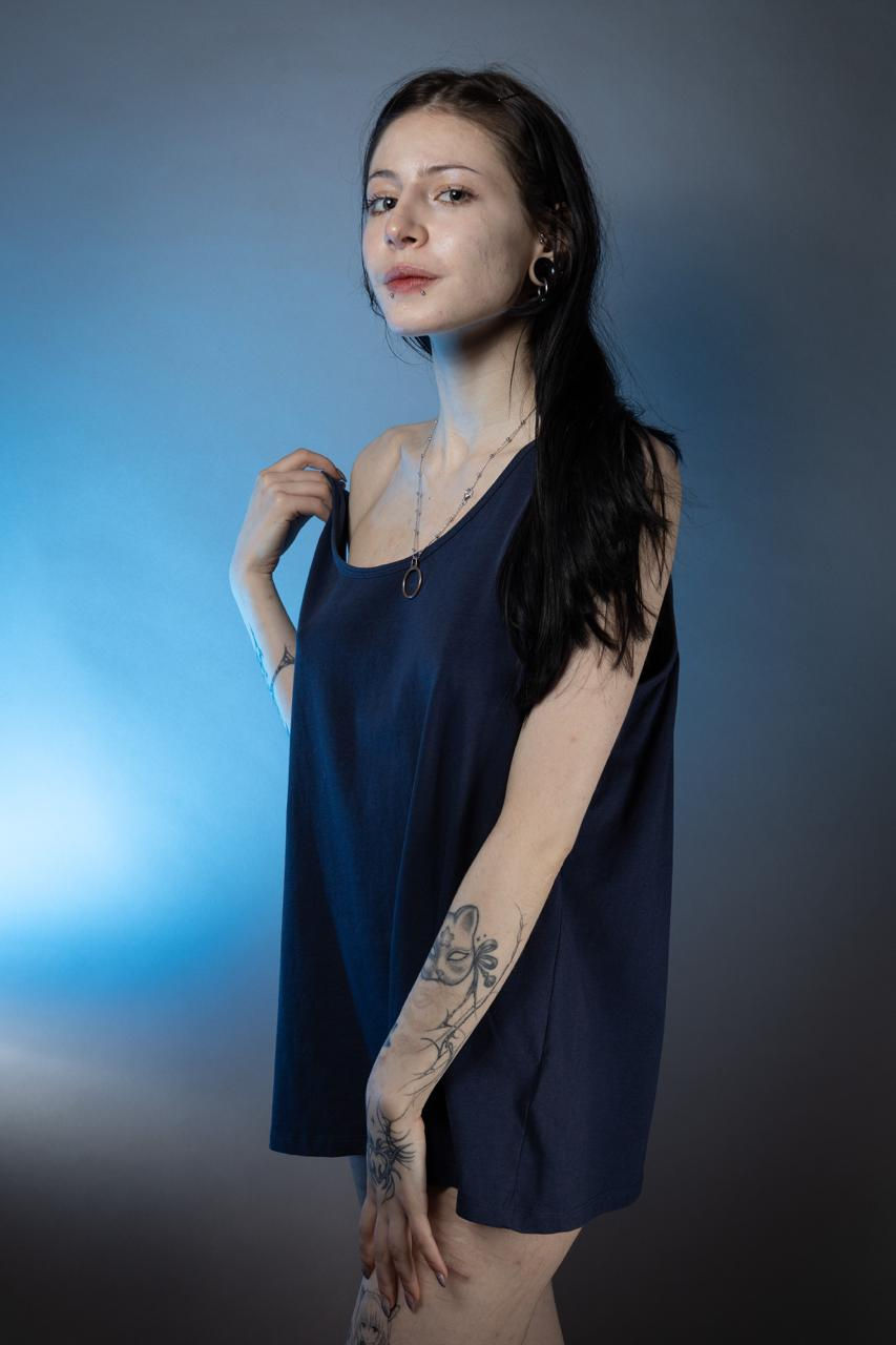 Alt text:

A person with long dark hair stands in front of a blue-to-gray gradient background, wearing an oversized dark sleeveless top that falls loosely over their torso. They have ear piercings, a lip piercing, and layered necklaces. One hand lightly holds the neckline of the shirt while the other rests by their thigh. Tattoos are visible on their forearm and upper arm, and they look at the camera with a neutral, slightly confident expression.
