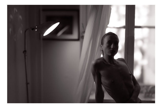 Black and white photograph of a person sitting by a window with natural light streaming in. A desk lamp is visible on the left side of the frame, and sheer curtains frame the window. The composition creates a contemplative, intimate atmosphere with soft lighting and shadows.