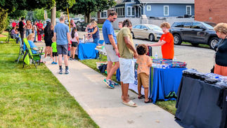 A lively crowd of neighbors walking past community booths and tables set up along the street.