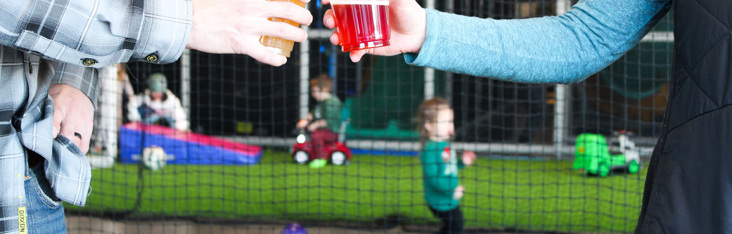 Two adults clinking drinks in an indoor play area with children playing