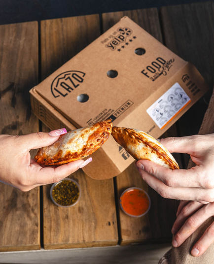Two hands holding empanadas, tips touching, with LAZO box and sauces.