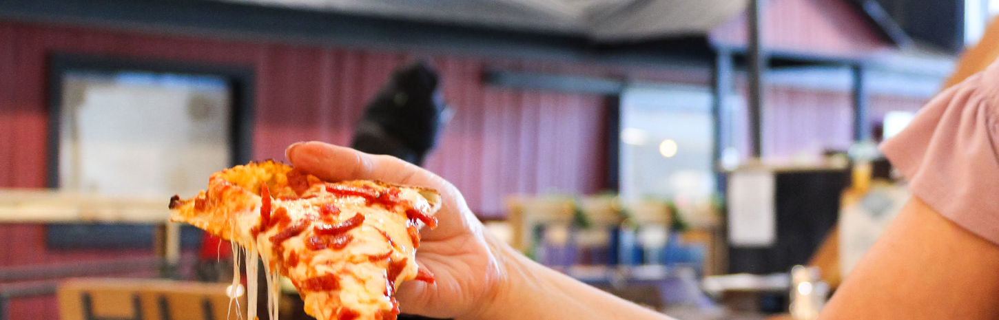 Hand lifting cheesy pizza slice, TV in background