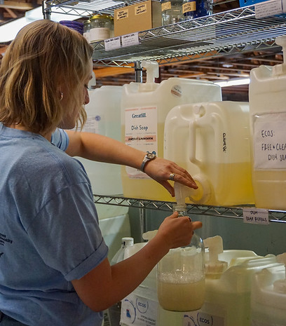 A woman with short blonde hair uses a bulk dish soap bin. She is turning the nozzle so the bulk container released liquid into a reusable container below.