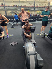 A man in athletic gear sits on a rowing machine in a bustling gym, smiling confidently