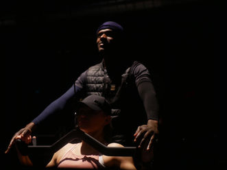A person in a dark-lit gym, wearing a cap, is being assisted by another person behind them during weight training