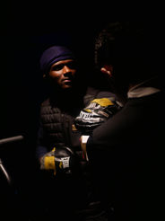 A man in a dark vest and blue beanie, wearing boxing gloves, is sparsely lit, facing another person holding boxing mitts