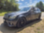 Black Mercedes-Benz station wagon parked on a sunny rural road beside lush greenery