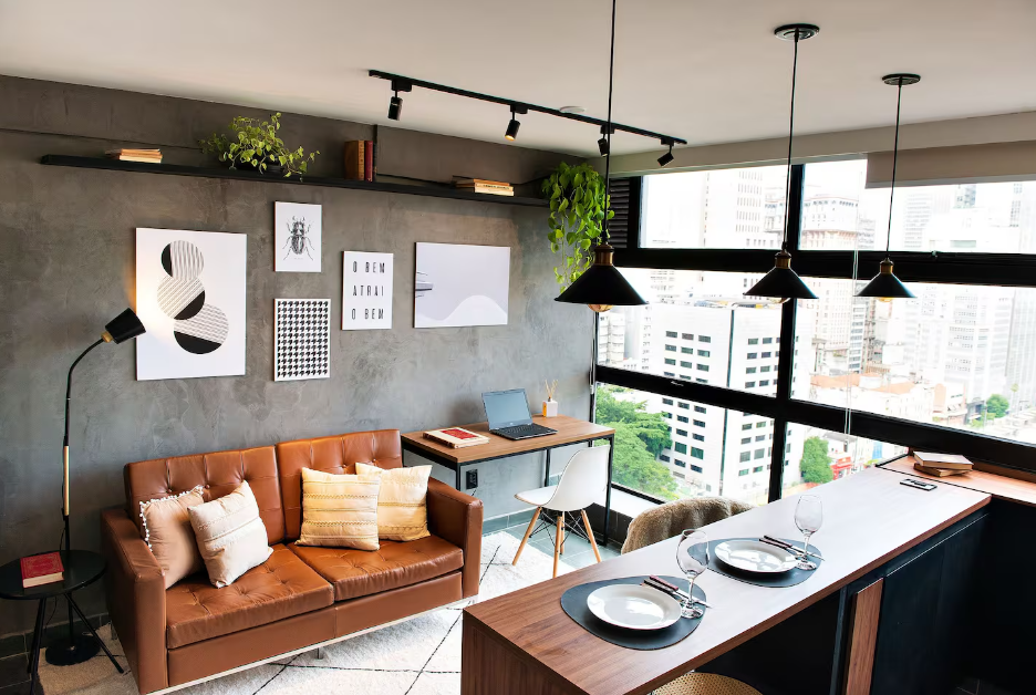 Sala com sofá caramelo, parede de concreto aparente e janelas amplas com iluminação natural.