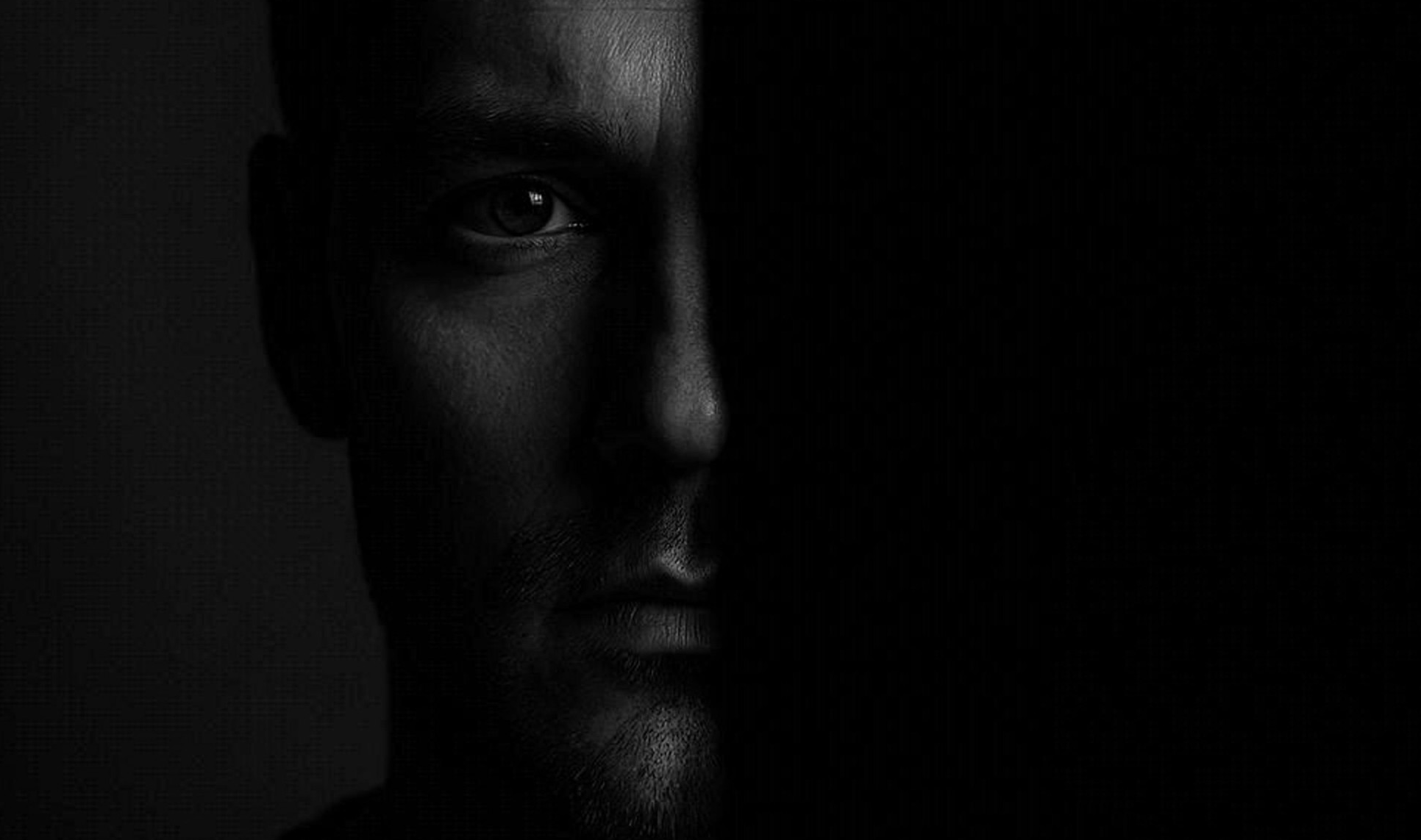 Half-lit man’s face in deep shadow, staring ahead in a moody black-and-white portrait.