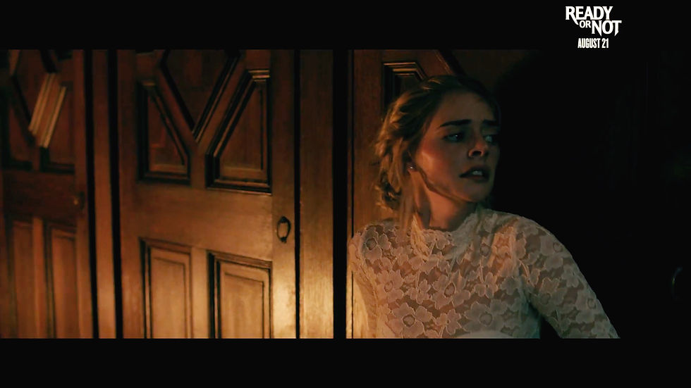 Woman leaning against a wood-paneled wall next to a wooden door. She's looking to the right. She has blonde, braided hair in an updo. She has fair skin. She is wearing a white lace wedding dress. The room is very dark and atmospheric. She looks scared.