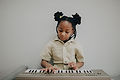 A young girl with braided hair tied up into pigtails sits in front of a keyboard piano and plays.