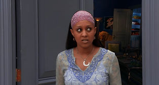A woman stands in a doorway with a confused look on her face. She is wearing a purple patterned head wrap. She's wearing a thin necklace with a crescent-moon pendant and a light blue-gray embroidered top.