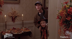 A young girl and a teenage boy peer around the corner of a room. There's a table to the left of the image with candles and Halloween decorations. The girl is wearing a witch’s hat.