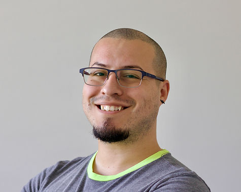 A photo of Thomas on a beige background. He is wearing glasses and a gray t-shirt with a neon yellow collar.