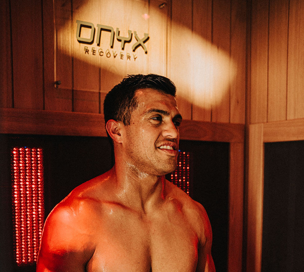 Shirtless man in a red-lit sauna, smiling and sweating, with an ONYX Recovery sign on the wood-paneled wall.