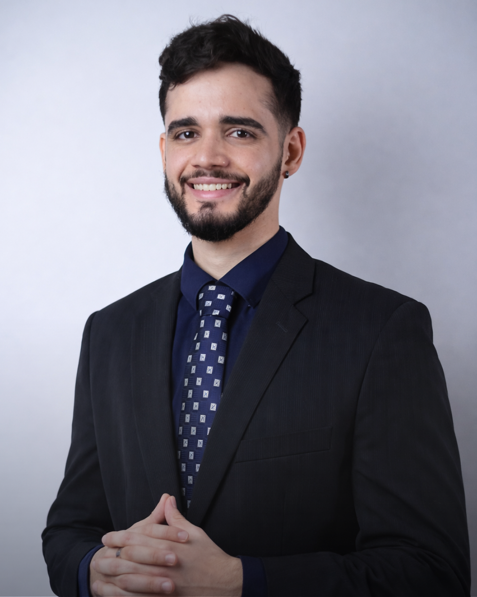 Imagem profissional de um homem com barba e cabelos pretos, roupa social, fundo branco neutro. Ele está com expressão serena