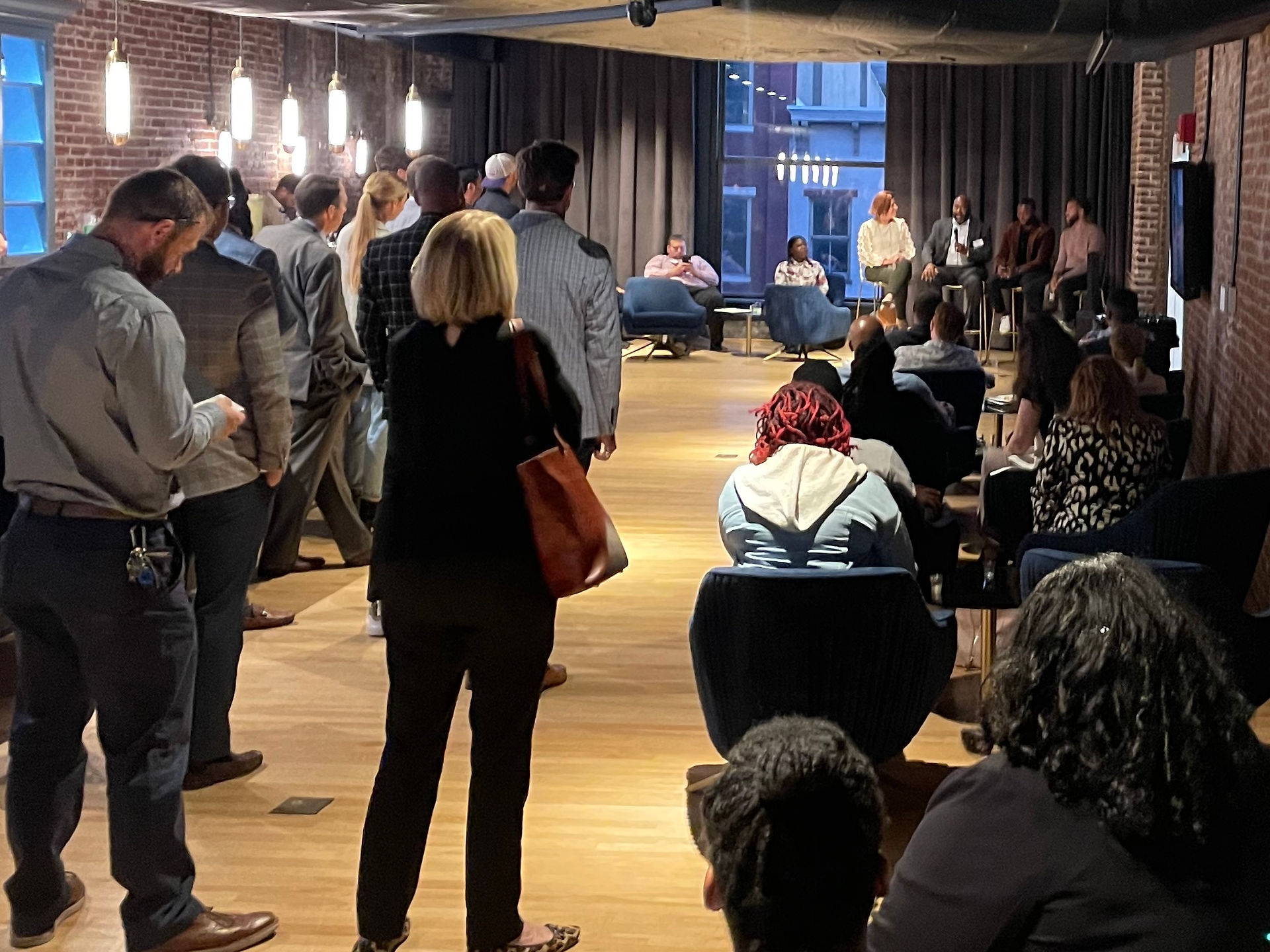 Crowd looks on in dimly lit speakeasy setting as four seated speakers converse.