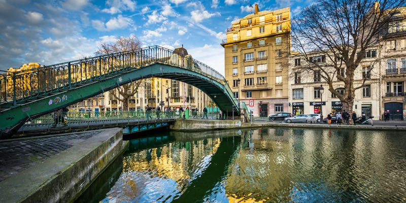 Bridge over canal Saint Martin in Paris with people walking, surrounded by historic buildings. Clear blue sky and calm water reflecting the vibrant city scene.