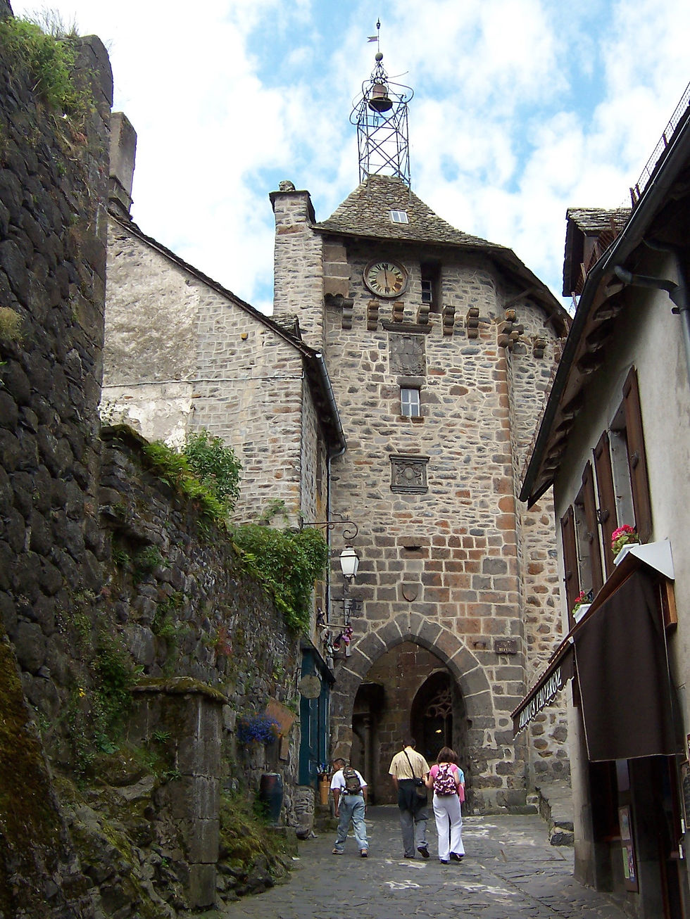 Three people walk down a cobblestone street toward a historic stone building with a clock tower. Sky is partly cloudy; plants line the walls.