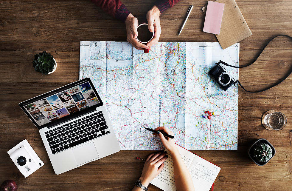 Overhead view of a table with a map, cameras, laptop, journals, and pens and papers scattered around. Hands are shown holding pens and pointing to the map. A cozy vibe indicating productive, focused trip planning with lots of decisions and details.