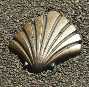 Bronze scallop shell embedded in textured pavement, symbolizing the Camino de Santiago. Sunlight highlights its glowing metal surface.