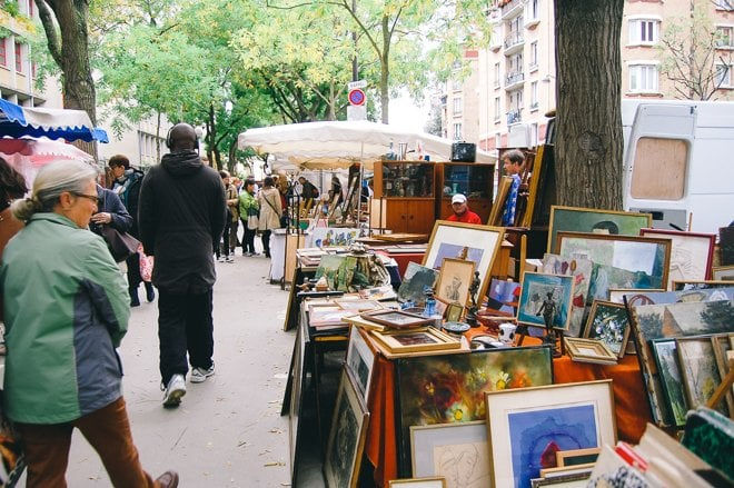 People browsing artwork at an outdoor flea market in Paris with colorful paintings. Trees line the street, and a white van is in the background.