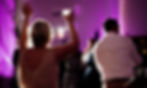 woman in sparkly dress holds drink and hand up in celebration on a dance floor lit with purple light.