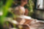A woman sits cross-legged on a bed with her hands at heart center, surrounded by natural light and indoor plants, representing a peaceful morning moment of mindfulness and intention.