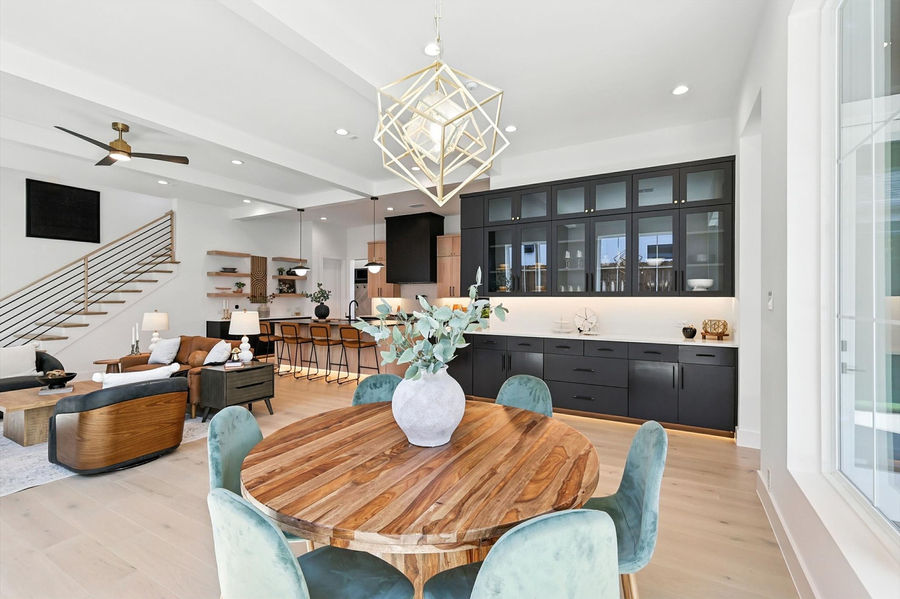 Staged open-concept dining and living area with a round wooden table, blue chairs, dark kitchen cabinets, and a modern light fixture.