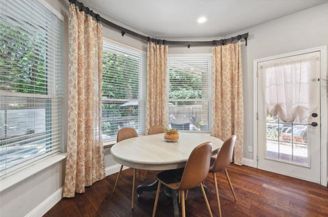 Bright and airy breakfast nook featuring a round white dining table paired with sleek modern chairs, surrounded by large windows dressed with elegant patterned drapes. The space is enhanced by warm hardwood floors, natural light, and a view of the inviting outdoor area, creating a cozy yet stylish spot for meals and gatherings.