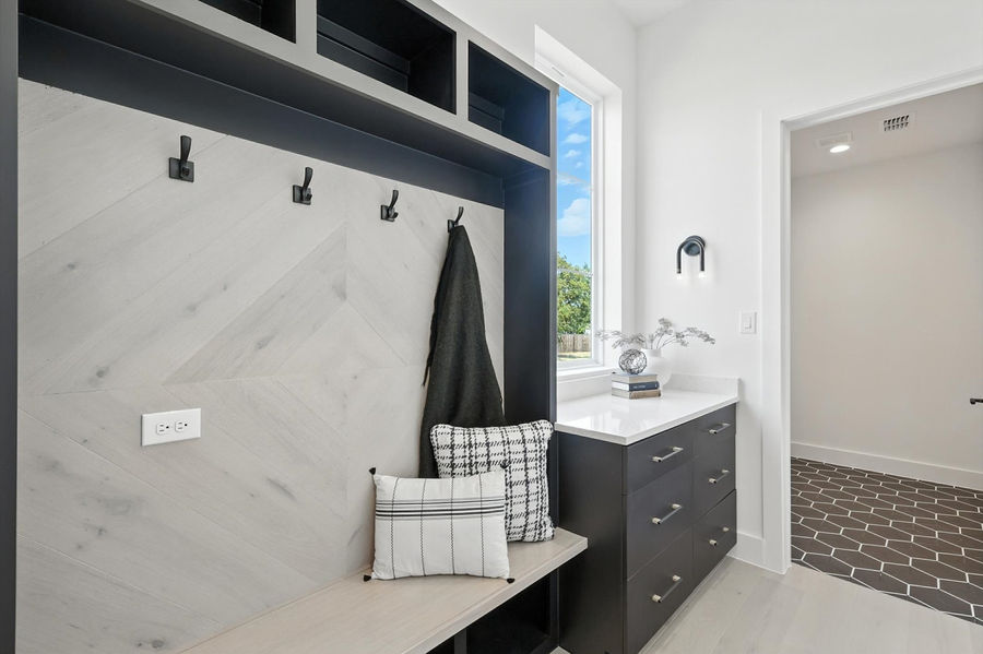 Close-up of a staged mudroom with a built-in bench, hooks, dark gray storage drawers with a white countertop and decor, and a glimpse into another room with patterned flooring.