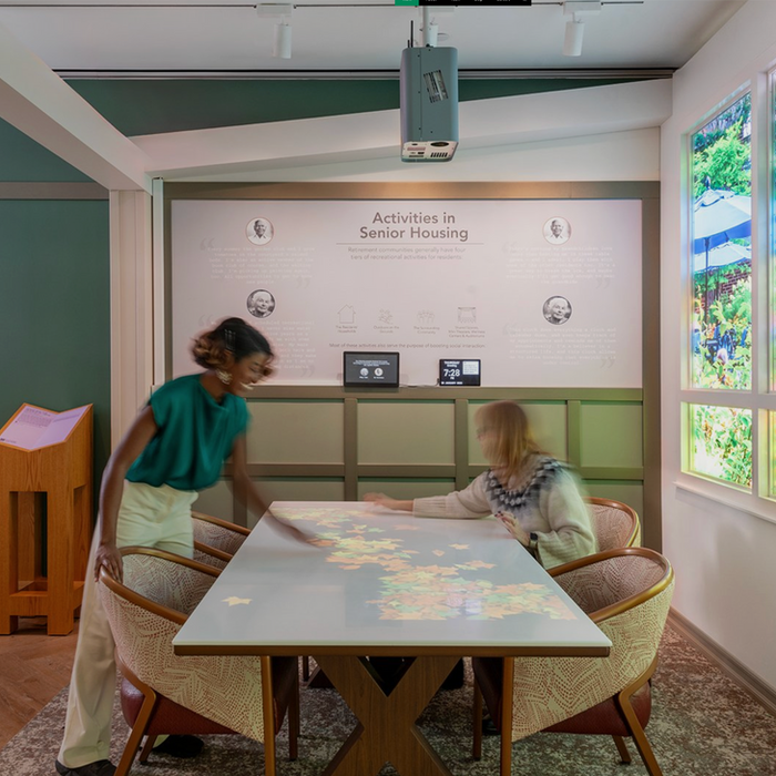 Two women interacting at a table in a thoughtfully designed senior living space, with a projected display of colorful shapes on the tabletop and informational wall graphics about activities in senior housing in the background.