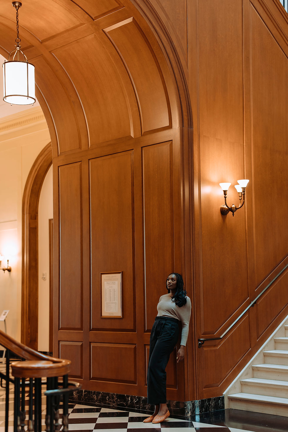 A woman (Tosin Odugbemi Hoskins) standing within a refined, classical interior with wood-panelled architectural space with arched detailing, marble flooring.