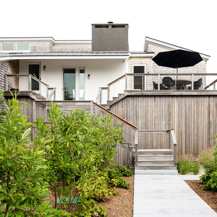 Modern coastal home exterior with weathered wood siding, elevated decks with metal railings, outdoor seating and an umbrella, and a landscaped walkway leading to the entrance.