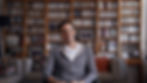 A man in a gray hoodie sits indoors, smiling at the camera with shelves and framed photos behind him.