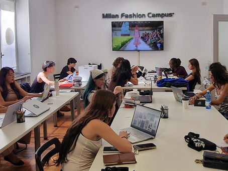 Students work on laptops in a Milan Fashion Campus classroom while a runway show plays on a wall screen.