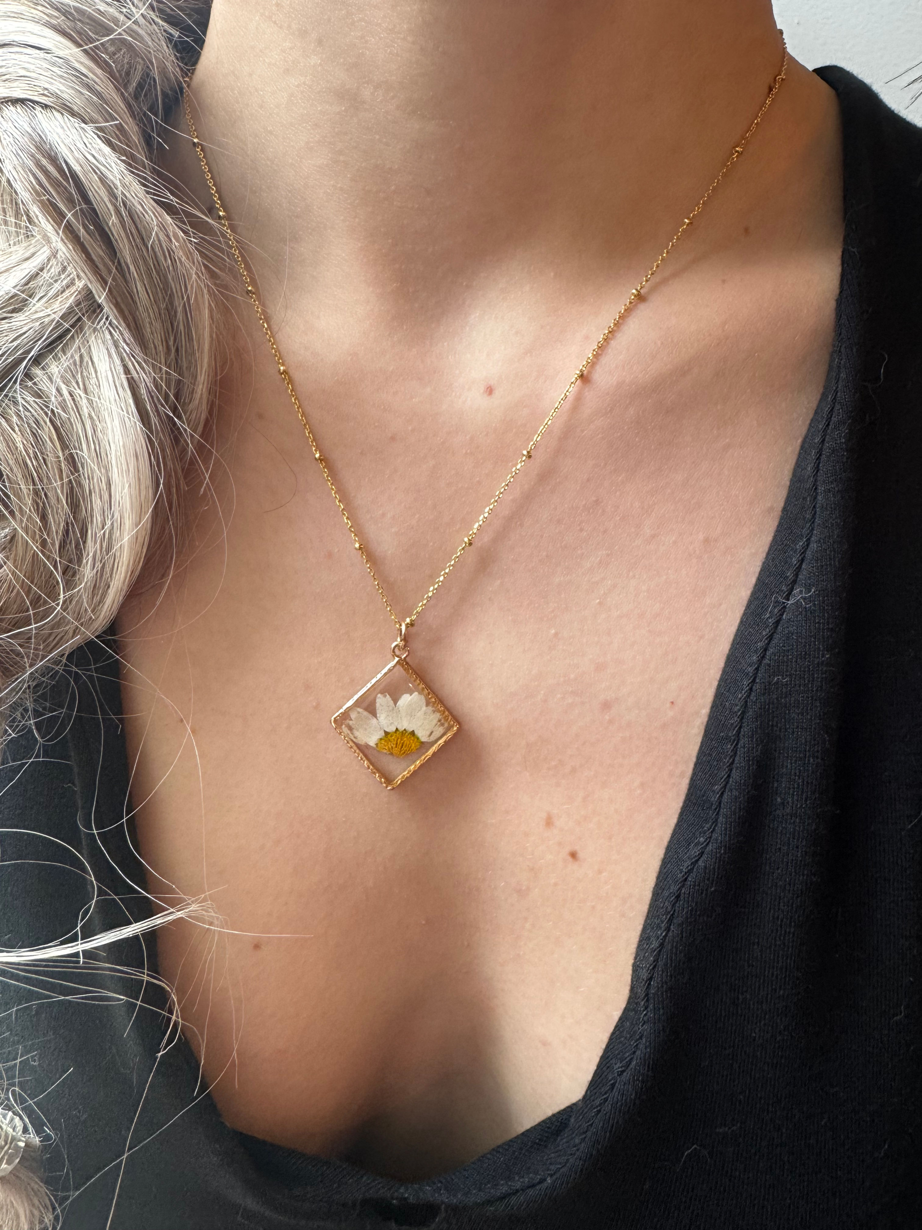 Woman wearing gold necklace with square dried flower pendant, black shirt.