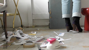 A film still featuring a close up shot of Sonia Boué's feet in high heeled blue suede t-bar shoes in a minimalist studio space. Collage materials are strewn on the floor and a tattered vintage suitcase can also be seen.
