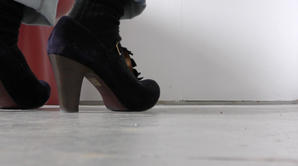 Film still of Sonia Boué's feet in her minimalist studio space. She is wearing high heeled blue suede t-bar shoes. There is a red stool in the shot. 