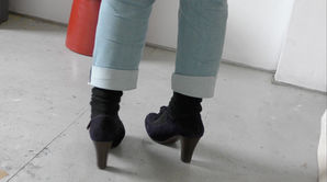 Film still of Sonia Boué's feet in her minimalist studio space. She is wearing high heeled blue suede t-bar shoes. There is a red stool in the shot.