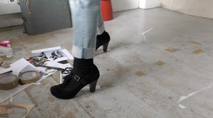 A film still featuring a close up shot of Sonia Boué's feet in high heeled blue suede t-bar shoes in a minimalist studio space. Collage materials are strewn on the floor and a tattered vintage suitcase can also be seen.