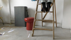 Film still of Sonia Boué's feet in her minimalist studio space on a wooden step ladder. She is wearing high heeled blue suede t-bar shoes. A red stool and tattered vintage suitcase can also be seen.
