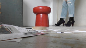 A film still featuring a close up shot of Sonia Boué's feet in high heeled blue suede t-bar shoes in a minimalist studio space. Collage materials are strewn on the floor and a tattered vintage suitcase can also be seen. 