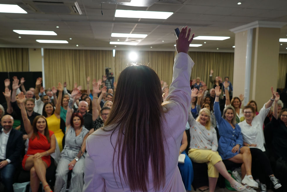 A person in a white blazer raises a hand facing an audience doing the same in a well-lit room. The mood is energetic and engaged. Mental fitness workshops