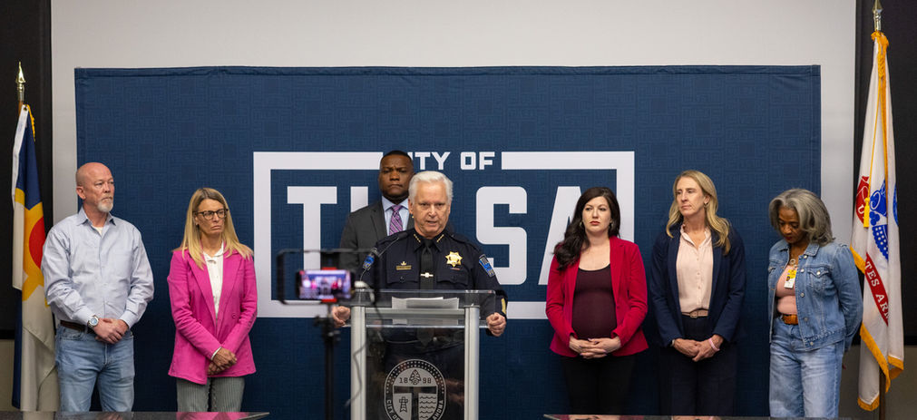 Councilor Bellis stands in the background with other Councilors and the Mayor while the police chief speaks at a press conference 