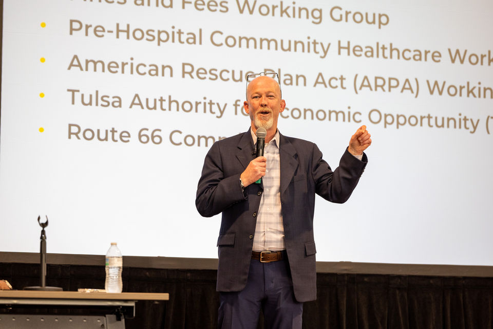 Councilor Christian Bengel standing in front of a large screen with a powerpoint presentation, holding a microphone and delivering a speech.